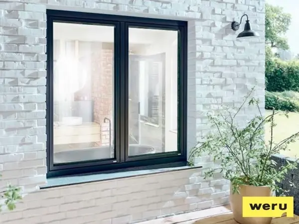 Modernes Fenster mit schwarzem Rahmen in weißer Klinkerfassade, daneben grüne Pflanze.